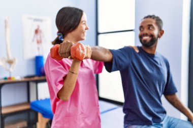 Man and woman wearing physiotherapist uniform having rehab session stretching arm holding dumbbell at physiotherpy clinic