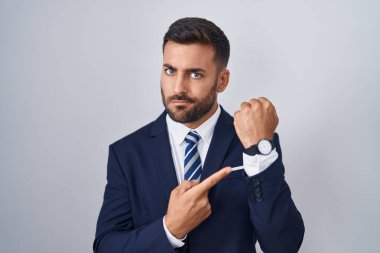 Handsome hispanic man wearing suit and tie in hurry pointing to watch time, impatience, looking at the camera with relaxed expression 