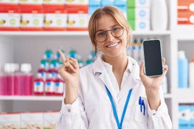 Young caucasian woman working at pharmacy drugstore showing smartphone screen smiling happy pointing with hand and finger to the side 