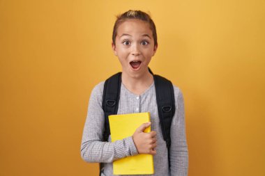 Little caucasian boy wearing student backpack and holding book scared and amazed with open mouth for surprise, disbelief face 