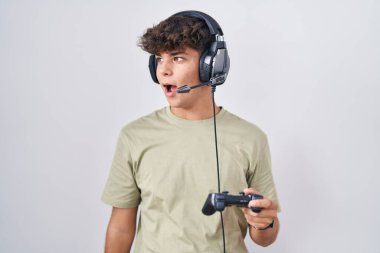 Hispanic teenager playing video game holding controller angry and mad screaming frustrated and furious, shouting with anger. rage and aggressive concept. 