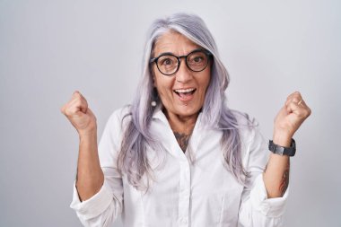 Middle age woman with tattoos wearing glasses standing over white background celebrating surprised and amazed for success with arms raised and open eyes. winner concept. 