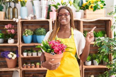 African american woman with braided hair working at florist shop holding plant smiling happy pointing with hand and finger to the side 