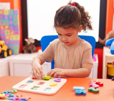 Adorable hispanic girl playing with maths puzzle game sitting on table at kindergarten