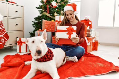 Kafkasyalı genç bir kadın boks hediyesini açıyor. Evde köpekle oturuyor. Noel ağacı yanında.