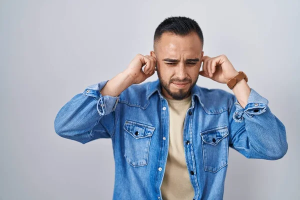 孤立した背景の上に立つ若いヒスパニック系の男は 大きな音楽のノイズのためにいらいらする表情で指で耳を覆う 聴覚障害の概念 — ストック写真
