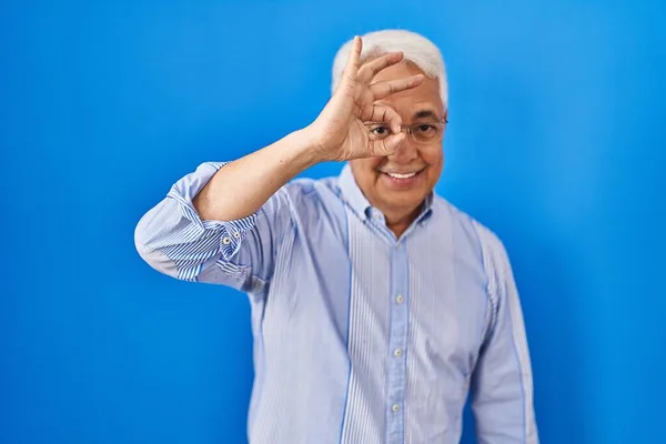 Hispanic senior man wearing glasses doing ok gesture with hand smiling, eye looking through fingers with happy face.