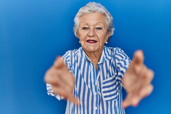 头发灰白的老妇人站在蓝色的背景上 看着镜头 张开双臂拥抱 欢快的表达拥抱幸福 — 图库照片