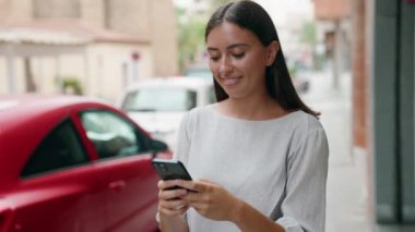 Genç, güzel, İspanyol bir kadın sokakta akıllı telefon kullanarak gülümsüyor.