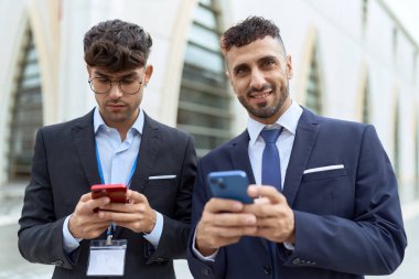 İki İspanyol işadamı sokakta akıllı telefon kullanarak gülümsüyor.