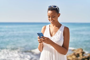Afrika kökenli Amerikalı bir kadın deniz kenarında akıllı telefon kullanarak gülümsüyor.