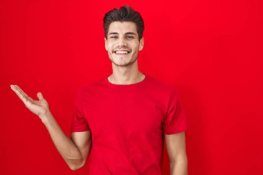 Kırmızı arka planda duran genç İspanyol adam mutlu bir şekilde sunum yapıyor ve avuç içiyle kameraya bakıyor.. 