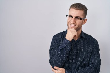 Young caucasian man standing over isolated background looking confident at the camera smiling with crossed arms and hand raised on chin. thinking positive. 