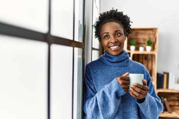 African american woman drinking coffee leaning on window at home