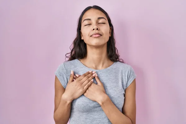 ピンクの背景にカジュアルなTシャツを着た若いブラジル人女性が目を閉じて胸に手で笑顔と顔に感謝のジェスチャー 健康の概念 — ストック写真
