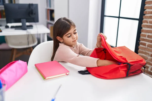 Adorable hispanic girl student sitting on table putting book on backpack at classroom