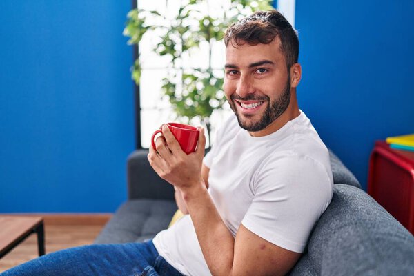 Young hispanic man drinking coffee sitting on sofa at home