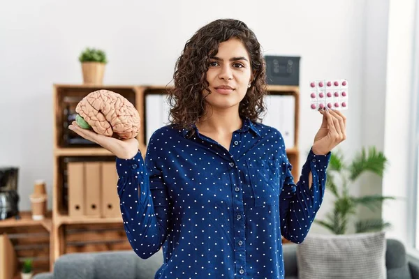 Young brunette woman holding brain and pills as mental health concept relaxed with serious expression on face. simple and natural looking at the camera.