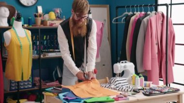 Young redhead man tailor smiling confident cutting cloth at clothing factory