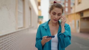 Young woman talking on smartphone holding credit card at street