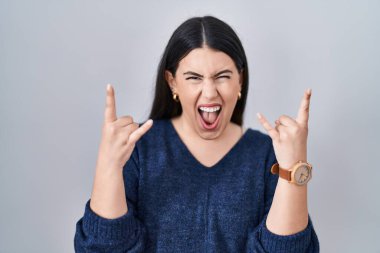 Genç esmer kadın izole edilmiş arka planda duruyor çılgın bir ifadeyle bağırıyor elleri havada rock sembolü yapıyor. Müzik yıldızı. ağır müzik konsepti. 