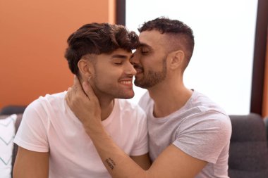 Two hispanic men couple sitting on sofa hugging each other and kissing at home