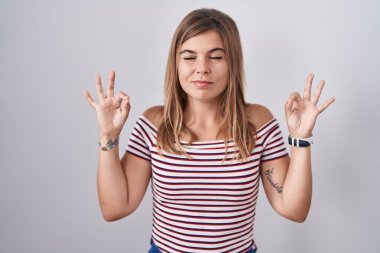 Tecrit edilmiş arka planda duran genç İspanyol kadın rahatlamış ve parmaklarıyla meditasyon hareketi yaparken gözleri kapalı gülümsüyor. yoga konsepti. 