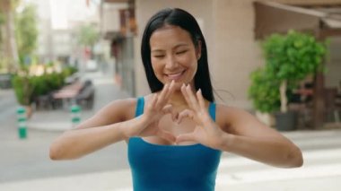 Young beautiful latin woman smiling confident doing heart gesture with hands at street