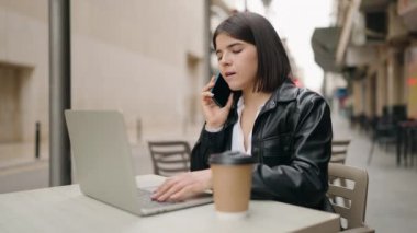 Young hispanic woman talking on the smartphone and using laptop at coffee shop terrace