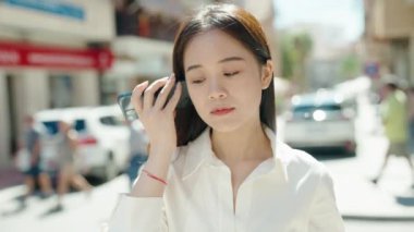 Young chinese woman smiling confident listening audio message by the smartphone at street