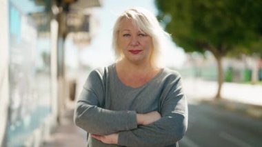 Middle age blonde woman with relaxed expression standing with arms crossed gesture at street