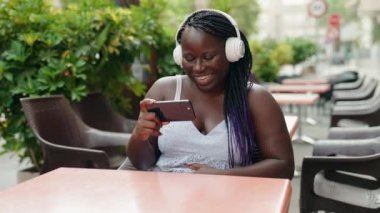 African american woman watching video on smartphone sitting on table at coffee shop terrace