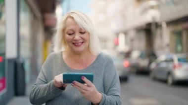 Orta yaşlı sarışın kadın gülümsüyor. Sokakta akıllı telefondan videolar izliyor.