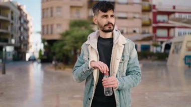 Young hispanic man standing with relaxed expression drinking water at street