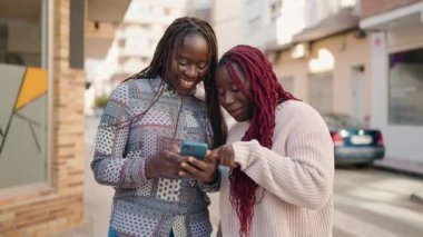 İki Afro-Amerikalı kadın sokakta akıllı telefon kullanarak gülümsüyor.