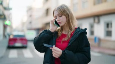 Young blonde girl talking on the smartphone and using credit card at street
