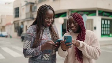 Two african american women smiling confident using smartphone at street