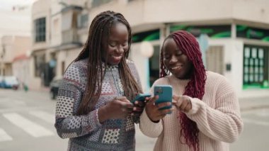 İki Afro-Amerikalı kadın sokakta akıllı telefon kullanarak gülümsüyor.