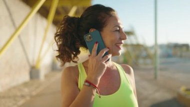 Orta yaşlı kadın spor kıyafeti giyiyor ve caddede akıllı telefondan konuşuyor.