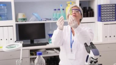 Orta yaşlı, gri saçlı, bilim adamı üniforması giyen bir kadın laboratuarda test tüplerini karşılaştırıyor.