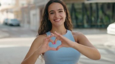 Genç, güzel, İspanyol bir kadın gülümsüyor. Kendine güveniyor, elleriyle kalp hareketi yapıyor.