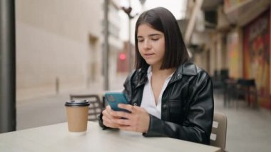 Young hispanic woman using smartphone drinking coffee at coffee shop terrace