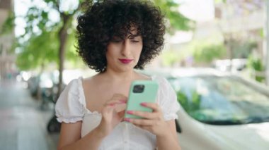 Orta Doğulu genç bir kadın sokakta akıllı telefon kullanarak gülümsüyor.