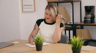 Young beautiful hispanic woman business worker listening voice message by smartphone writing on notebook at office