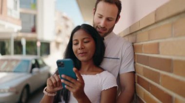 Man and woman interracial couple hugging each other using smartphone at street