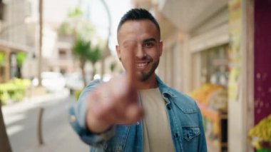 Young hispanic man smiling confident doing coming gesture with hand at street