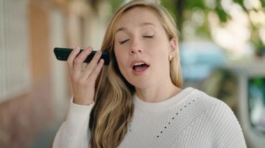 Akıllı telefondan sesli mesaj dinleyen genç sarışın kadın. Sokakta ciddi bir ifadesi var.