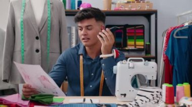 Genç İspanyol terzi akıllı telefondan konuşuyor giyim fabrikasında kıyafet tasarımı yapıyor.