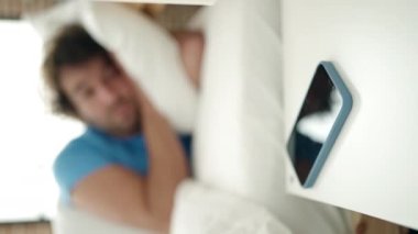 Young hispanic man lying on bed turning off smartphone alarm sleeping at bedroom