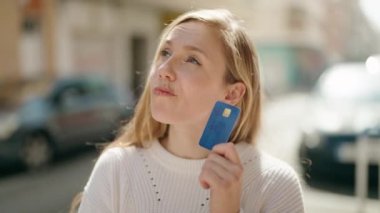 Young blonde woman holding credit card with doubt expression at street